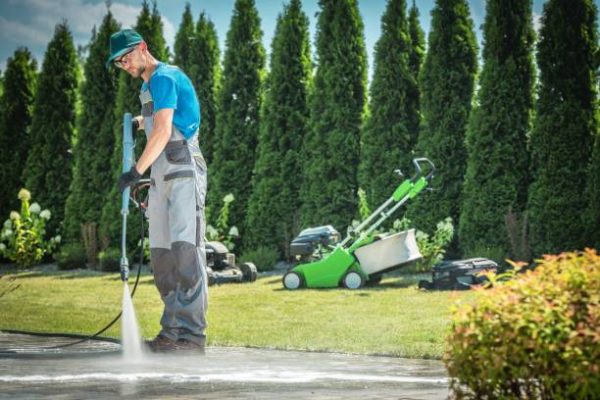 Caucasian Men in his 30s Cleaning Garden Paths and Driveway Using Professional Pressure Washer. Garden Equipment in the Background.